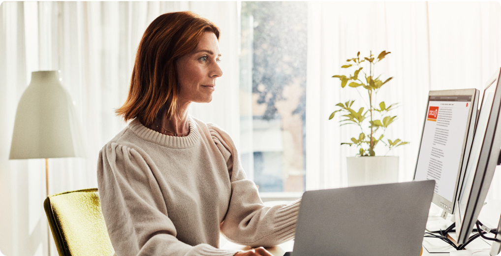 Woman working on a laptop in a bright home office, with a monitor and a plant nearby.
